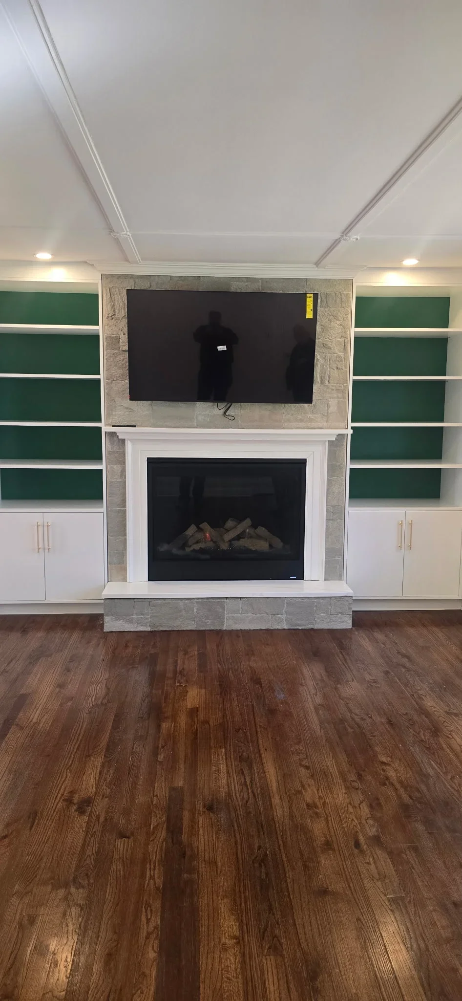 Stone fireplace with white mantel, gas insert, mounted TV, green shelving, white cabinets, hardwood floor